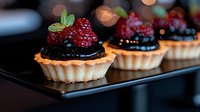 Mini tarts with chocolate ganache filling topped with fresh raspberries and mint leaves on black serving plate. Elegant dessert with blurred bokeh background.
