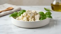 Fresh mozzarella cheese cubes with basil leaves and black pepper in white bowl on marble surface for Mediterranean cooking and healthy eating.