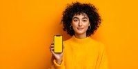 Young woman with curly hair in yellow sweater showing smartphone with blank screen against vibrant orange background for mockup design.