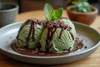 Green tea ice cream scoops with chocolate sauce drizzle and fresh mint garnish on ceramic plate. Matcha dessert for restaurant menu design.