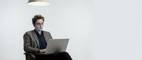 Young Caucasian businessman in brown suit working on laptop computer in modern minimalist office space with pendant lighting for corporate presentations.