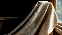 Mysterious figure draped in textured beige fabric sits by window in dark moody setting, creating dramatic contrast between light and shadow. Artistic portrait concept.
