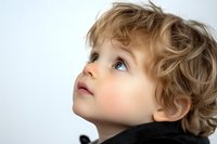 Young Caucasian child with curly blonde hair and blue eyes looking up with wonder and curiosity against white background, profile view showing innocent expression.