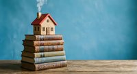 Small wooden house model sitting on stack of architecture books against blue wall background for real estate education and home design concepts.