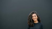 Young woman with long curly dark hair and warm smile wearing casual dark sweater against neutral gray background. Portrait for business, lifestyle, and professional use.