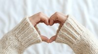 Hands in cozy knit sweater sleeves forming a heart shape against soft white background, symbolizing love, warmth, and care.