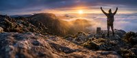 Young man with arms raised celebrating success on mountain peak at sunrise, symbolizing achievement and personal growth for motivational and business concepts.