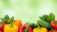 Fresh organic vegetables including colorful bell peppers, tomatoes, cucumbers and leafy greens arranged on green blurred background for healthy eating concepts.