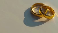 Two golden wedding bands positioned together on a neutral surface, casting soft shadows. Suitable for wedding invitations, engagement announcements, and bridal marketing materials.