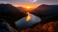Dramatic sunset over meandering river valley between mountains, golden sunburst creates light rays through clouds, reflection on calm water surface, autumn landscape view.