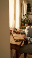 Cozy morning scene with person writing in journal at wooden table by sunny window with coffee cup and houseplants in warm home interior.