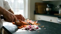 Chef hands chopping fresh vegetables on cutting board in modern kitchen. Professional food preparation with carrots and onions for healthy cooking.