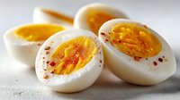 Hard-boiled eggs cut in half showing golden yolks sprinkled with paprika spice on white surface for healthy breakfast cooking and meal preparation.