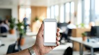 Hand holding smartphone with blank white screen in modern office environment with blurred colleagues and workspace in background.