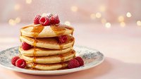 Stack of golden pancakes topped with fresh raspberries and maple syrup on decorative plate with warm bokeh lights creating cozy breakfast atmosphere.