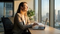 Happy professional businesswoman working on laptop in modern office with city view during golden hour sunlight.