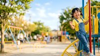 Young East Asian girl in denim overalls playing on yellow playground equipment at sunny park with trees and swings in background.