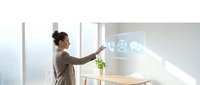 Woman in business casual attire interacting with holographic interface displaying digital icons in modern office workspace with natural light and potted plant.