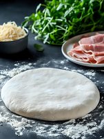 Fresh pizza dough on dark background with prosciutto, grated mozzarella and arugula leaves. Raw ingredients for traditional Italian cuisine preparation.