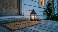 Cozy front porch at dusk with a welcome mat, glowing lantern, and small pumpkin beside a rustic wooden door for autumn home decor.