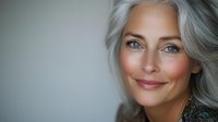Elegant mature woman with silver gray hair and bright blue eyes smiling confidently against neutral background, showcasing natural aging and beauty.
