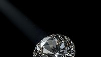 Brilliant cut diamond with sparkling facets on dark background with dramatic lighting beam creating luxury jewelry advertisement composition.