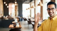 Young man with dark hair and glasses wearing a bright yellow shirt, waving hello in a modern office or coworking space with warm lighting and blurred colleagues in background.
