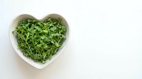 Fresh green arugula leaves in heart-shaped white ceramic bowl on clean background for healthy eating and cooking concepts.