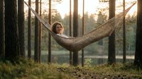 Young woman relaxing in hammock among tall trees during golden hour sunset, peaceful forest camping and outdoor recreation lifestyle moment.