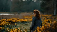 Young woman with curly red hair in cozy sweater standing in golden flower field during sunset, peaceful outdoor lifestyle portrait for wellness content.