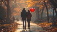 Romantic couple walking in autumn park holding red heart balloon against golden foliage background on misty morning, rear view on alley path.