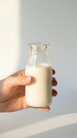 Hand holding glass bottle filled with fresh milk against neutral background with natural sunlight, suitable for dairy product advertising and food packaging design.