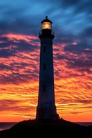 Majestic lighthouse silhouetted against dramatic orange and purple sunset sky with illuminated beacon light glowing through stormy clouds over ocean horizon.