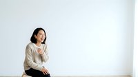 Young East Asian woman with shoulder-length dark hair wearing a beige cardigan, sitting and laughing with hand near chest against white background for lifestyle and portrait use.