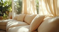 Sunlit cream-colored sofa with soft cushions near window with flowing beige curtains creating cozy living room atmosphere. Natural morning light casting warm glow.