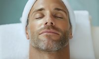 Caucasian man with stubble beard relaxing during facial treatment at spa, wearing white headband with eyes closed, expressing serenity and wellness.