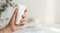 Unisex hand holding blank white cosmetic tube against neutral background with blurred greenery, suitable for skincare and beauty product packaging mockups.