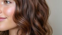 Close up portrait showing half face with wavy brown hair, natural makeup, and glowing skin against neutral background. Professional beauty and haircare concept.