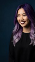 Young Asian woman with vibrant purple hair and dark lipstick smiling confidently against dark blue background for beauty and fashion concepts.