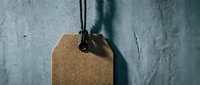 Blank brown kraft paper price tag hanging on black string against textured white wall with dramatic shadow lighting for retail and commercial use.