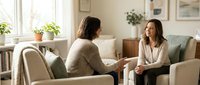 Two women having friendly conversation in bright modern living room with natural light from large windows and green plants creating warm atmosphere for counseling.