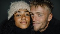 Young interracial couple in winter clothing embracing intimately against dark background, woman wearing knit beanie, cozy romantic portrait for lifestyle content.