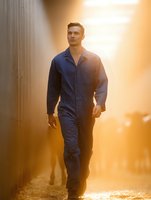 Caucasian male worker in blue coveralls walking confidently through industrial setting at sunset, dramatic golden backlight creates atmospheric mood for workplace safety concepts.