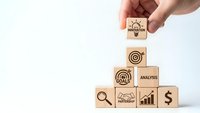 Hand placing wooden blocks with business icons forming pyramid structure representing strategic planning and growth concepts for corporate success.