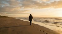 Solitary man walking along empty beach at golden hour sunset with dramatic cloudy sky and ocean waves, peaceful coastal scene for wellness and travel concepts.