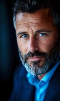 Close-up portrait of mature Caucasian businessman with gray beard and striking blue eyes wearing navy suit and blue shirt against dark background.