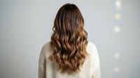 Young woman with long wavy brown hair wearing cream knit sweater from behind against white wall with soft lighting for hair care and beauty concepts.