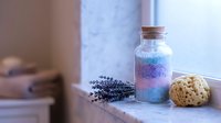 Colorful layered bath salts in glass jar with cork lid, dried lavender sprigs and natural sponge on bathroom windowsill for spa relaxation and self-care.