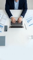 Professional businessman in navy suit typing on laptop at white desk with financial reports and charts for business analysis and corporate work.