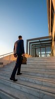 Caucasian businessman in navy suit carrying briefcase climbing marble stairs toward modern office building entrance at sunset.
